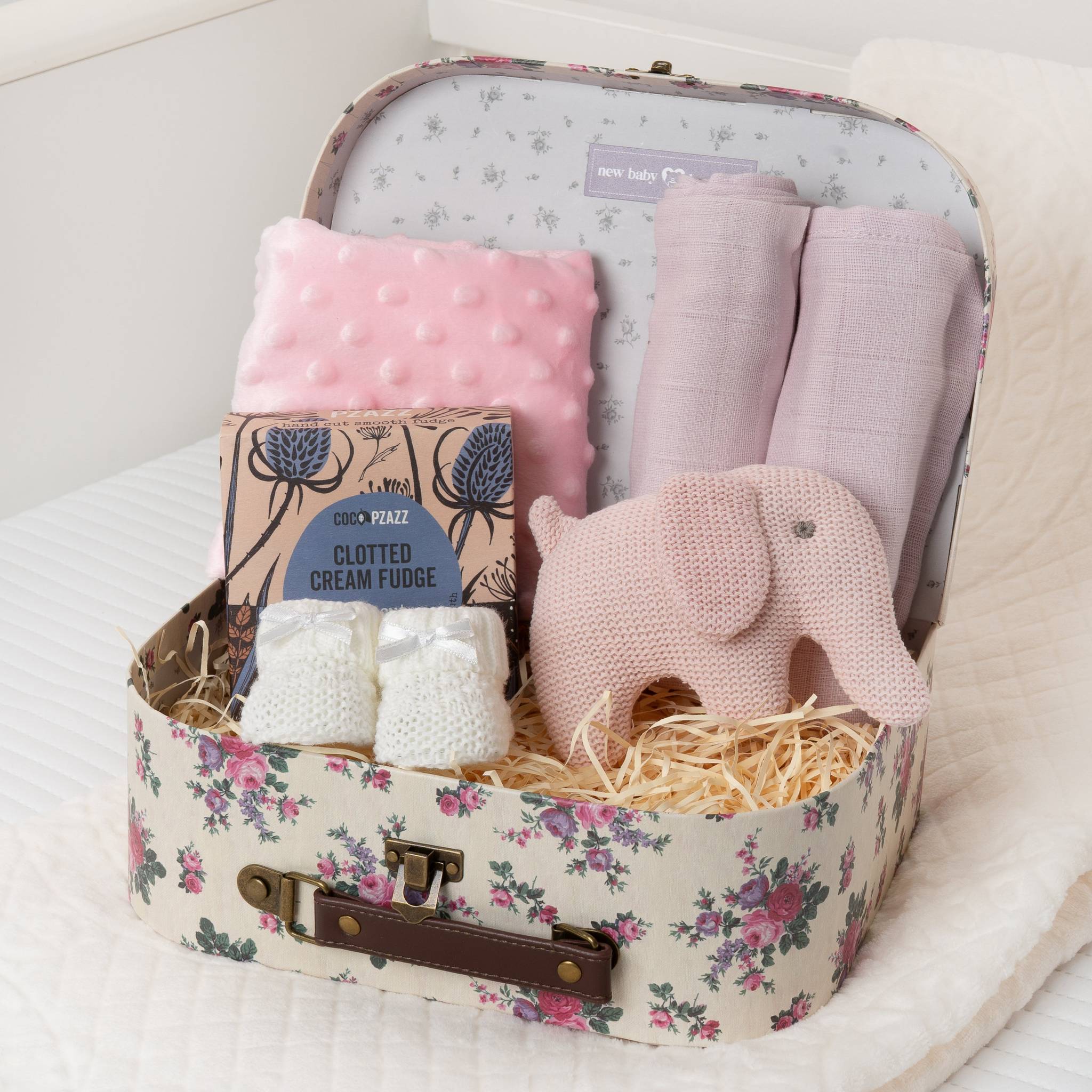 Floral-patterned suitcase filled with baby items including a pink plush elephant, pink blanket, and white fudge. Main image